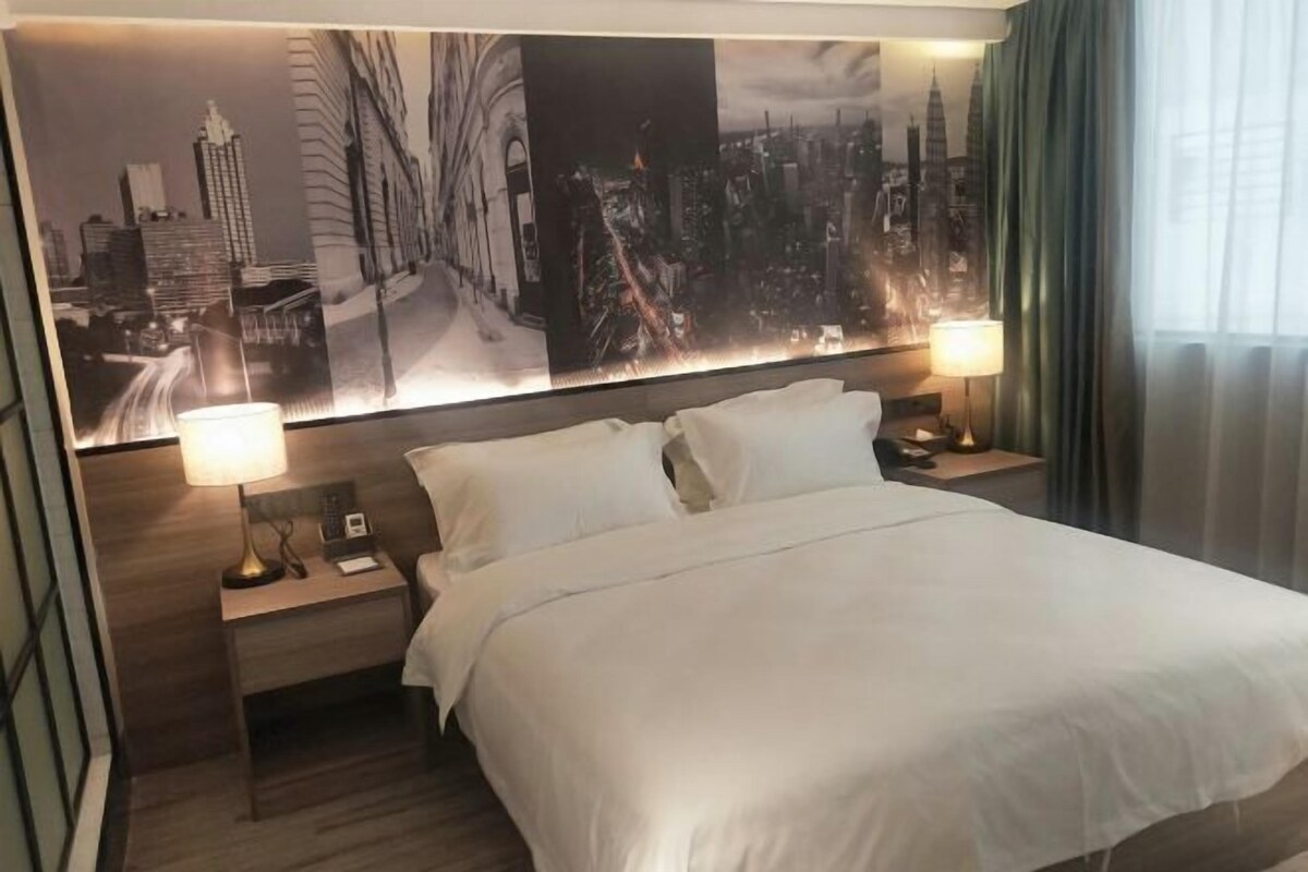 A hotel room with a large bed, two bedside tables with lamps, a cityscape mural on the wall, and a view of the city through the window.