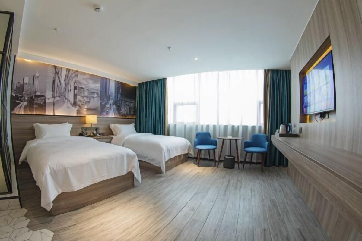 A hotel room with two beds, a desk, two chairs, a TV, and a cityscape painting on the wall.