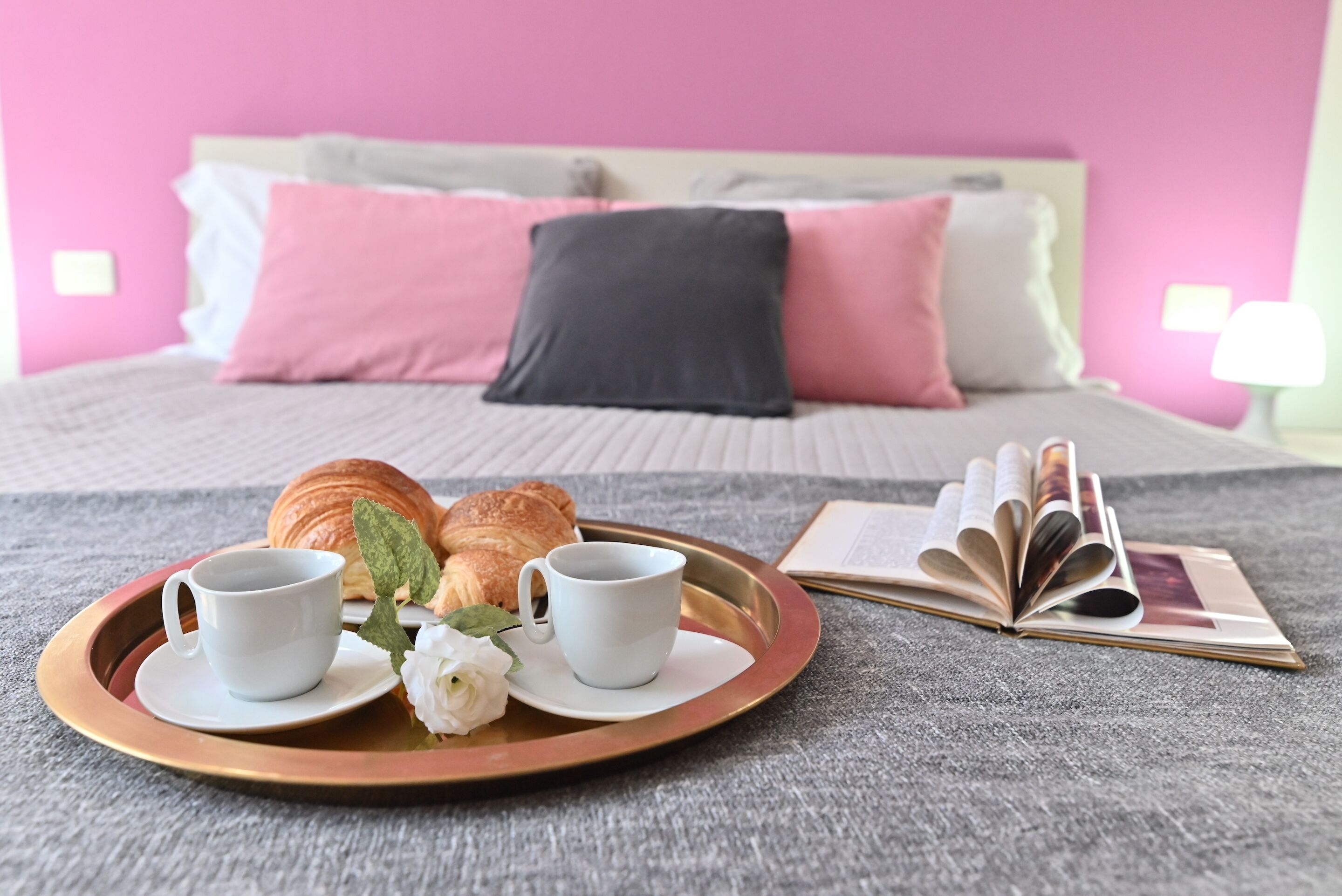 A breakfast tray with croissants and coffee cups on a bed with pink and gray bedding.