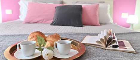 Bandeja de desayuno con cruasanes y tazas de café sobre una cama con ropa de cama rosa y gris.