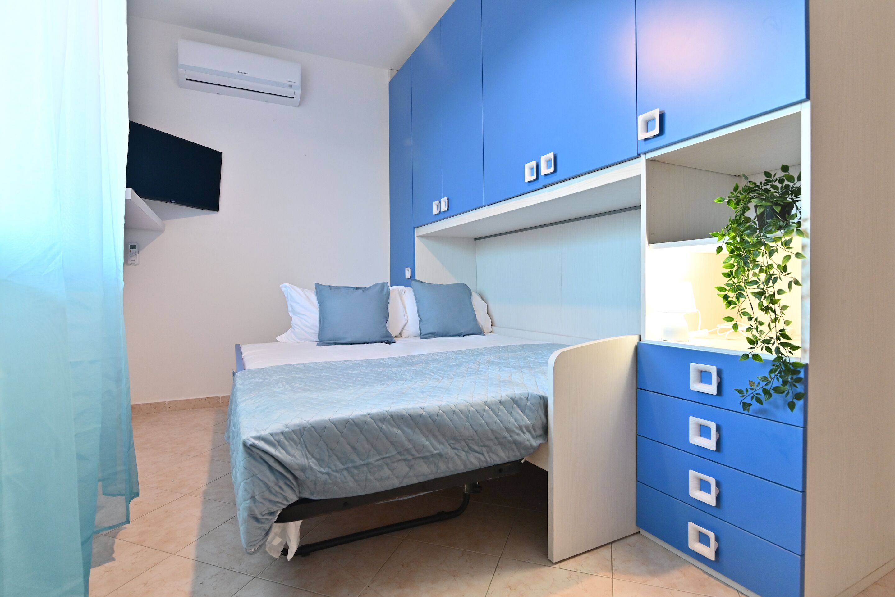 A bedroom with a bed, a blue wardrobe, a wall-mounted TV, and an air conditioner.