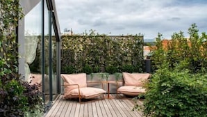 A wooden deck with two cushioned chairs and a table, surrounded by greenery and a glass wall. - Sublime Lisboa (Lisbon)