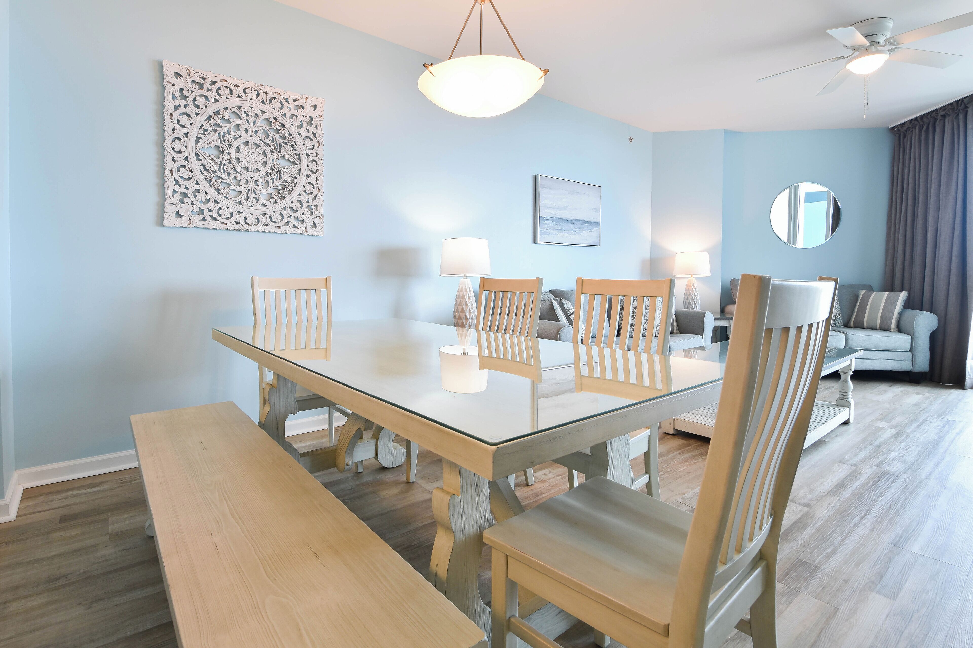 A dining area with a glass-top table, wooden chairs, and a bench. There is a ceiling fan, a decorative wall piece, and a sofa in the background.