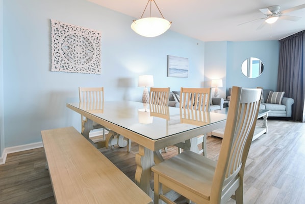 A dining area with a glass-top table, wooden chairs, and a bench. There is a ceiling fan, a decorative wall piece, and a sofa in the background.