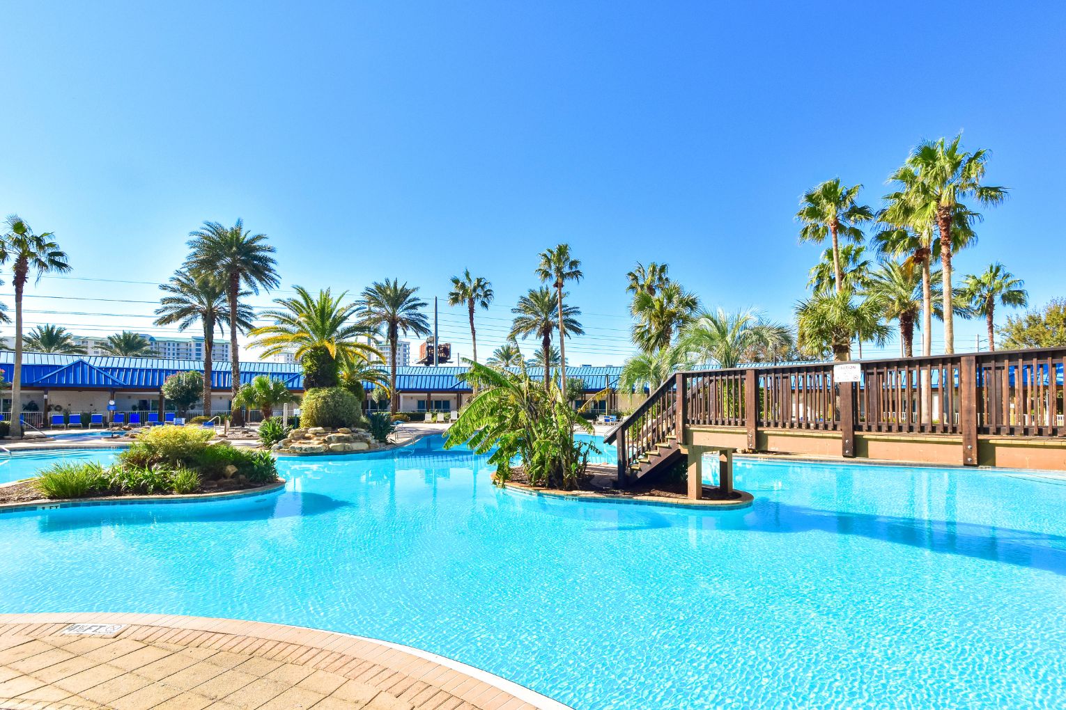 A swimming pool with a wooden deck, palm trees, and a building with blue awnings.