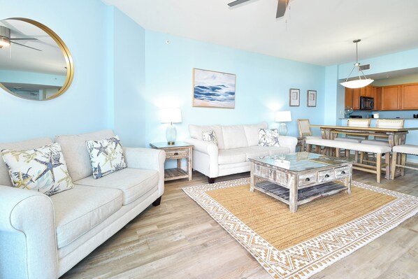 Uma sala de estar com parede azul clara, um espelho redondo, uma mesa de centro, um sofá com almofadas decorativas, um tapete e vista para a cozinha.