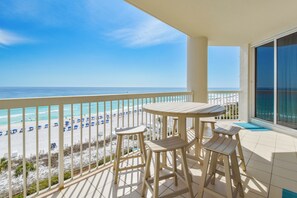 Un balcón con una mesa y taburetes, con vista a una playa con sombrillas y un cielo despejado.