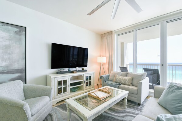 A modern living room with a flat-screen TV, a sofa, a coffee table, and a view of the ocean through large windows.