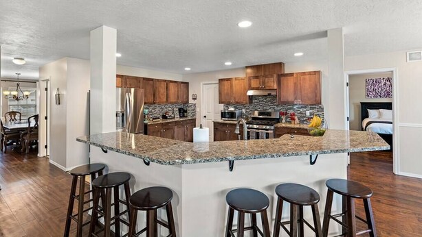 A modern kitchen with granite countertops, wooden cabinets, and a central island with bar stools. - The Tangle by Host Boise - 4500sqft Boise Home| 9 Beds & Dual Master| 85" TV (Boise)