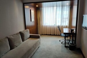 Una habitación de hotel con sofá, escritorio y un ventanal con cortinas translúcidas.