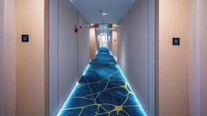 Un couloir d’hôtel avec un tapis bleu orné d’un motif géométrique jaune, des murs de couleur claire et un plafond doté d’un éclairage encastré.