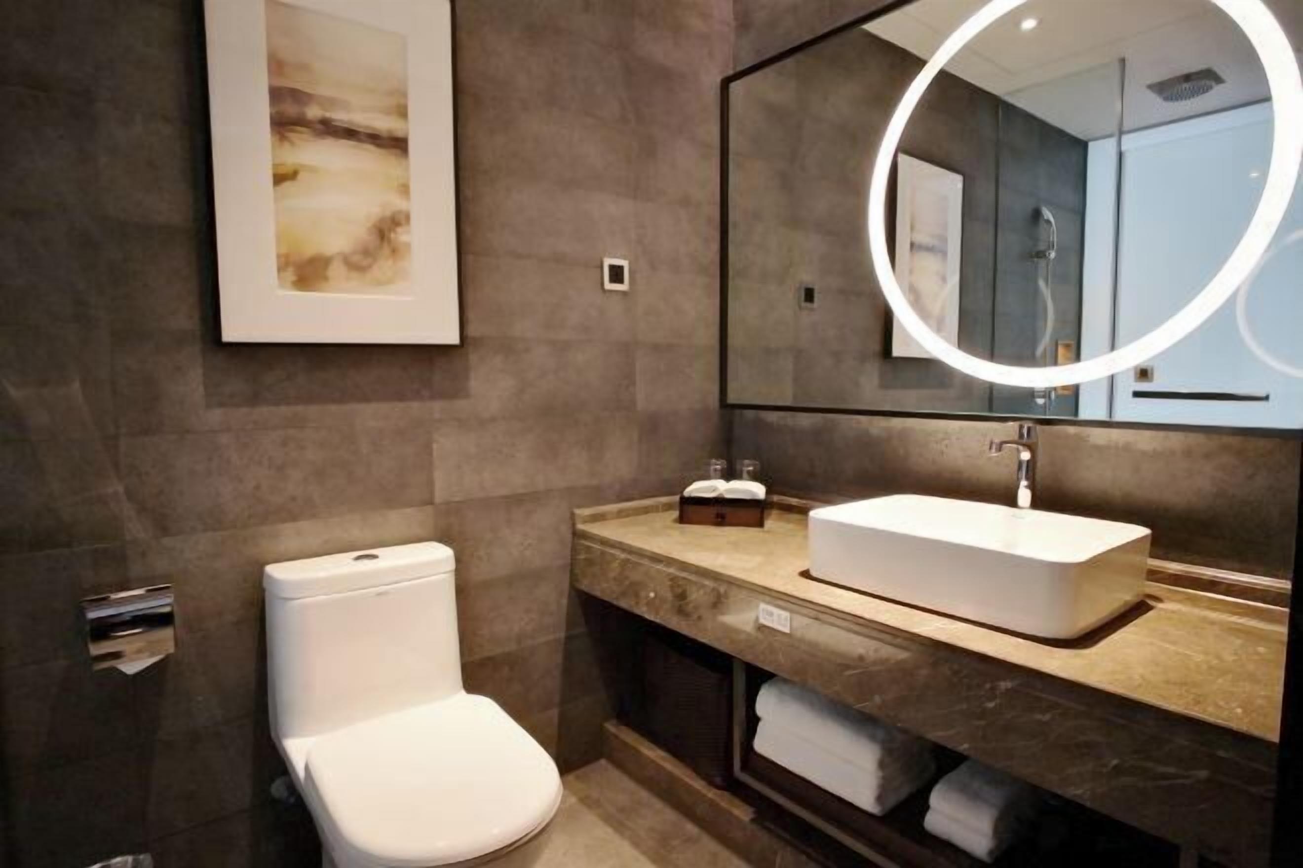 A modern bathroom with a white toilet, a rectangular sink, and a large mirror with a circular light.