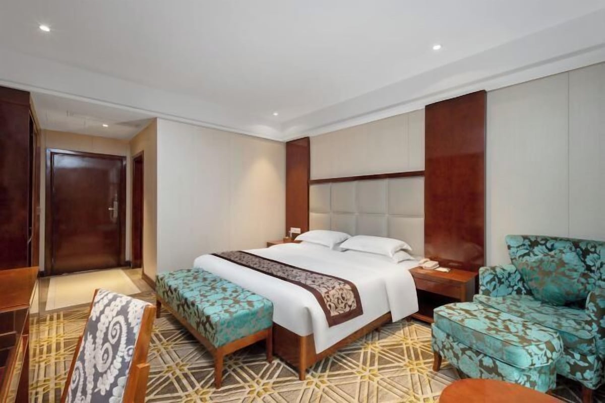 A hotel room with a large bed, a wooden headboard, a nightstand, a blue patterned armchair, and a patterned bench.