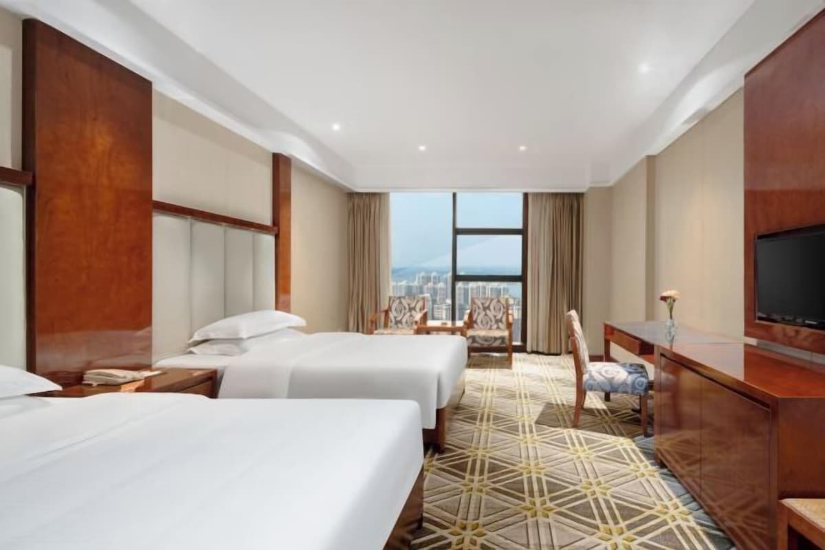 A hotel room with two beds, a large window with a city view, a television, and a desk with a chair.