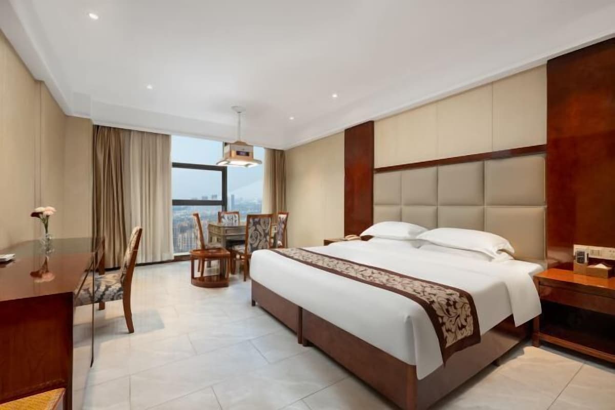 A hotel room with a large bed, a desk, and a chair. There is a window with curtains, a chandelier, and a view of the city.