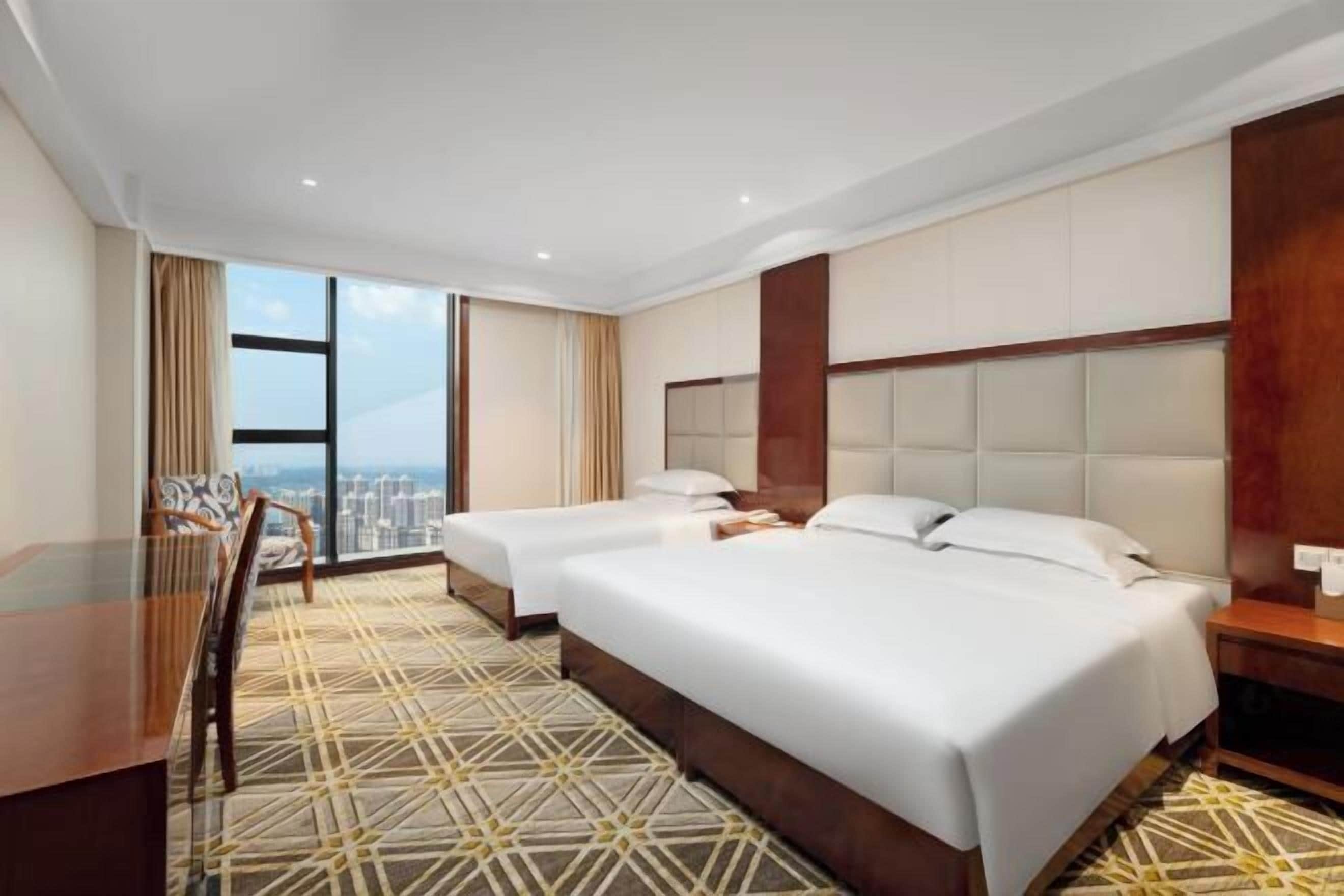 A hotel room with two beds, a desk, and a view of the city.