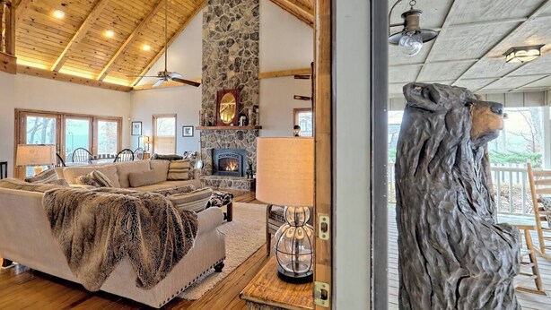 A cozy living room with a stone fireplace, wooden ceiling beams, and a large window overlooking a deck. - The Gathering Place by Chatuge Home Concierge | Mountain Views · Lake Chatuge Views (Hiawassee)