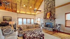 A spacious living room with a stone fireplace, wooden ceiling beams, and a large window overlooking a scenic landscape. - The Gathering Place by Chatuge Home Concierge | Mountain Views · Lake Chatuge Views (Hiawassee)