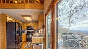 A kitchen with wooden cabinets, stainless steel appliances, and a large window overlooking a snowy deck. - The Gathering Place by Chatuge Home Concierge | Mountain Views · Lake Chatuge Views (Hiawassee)