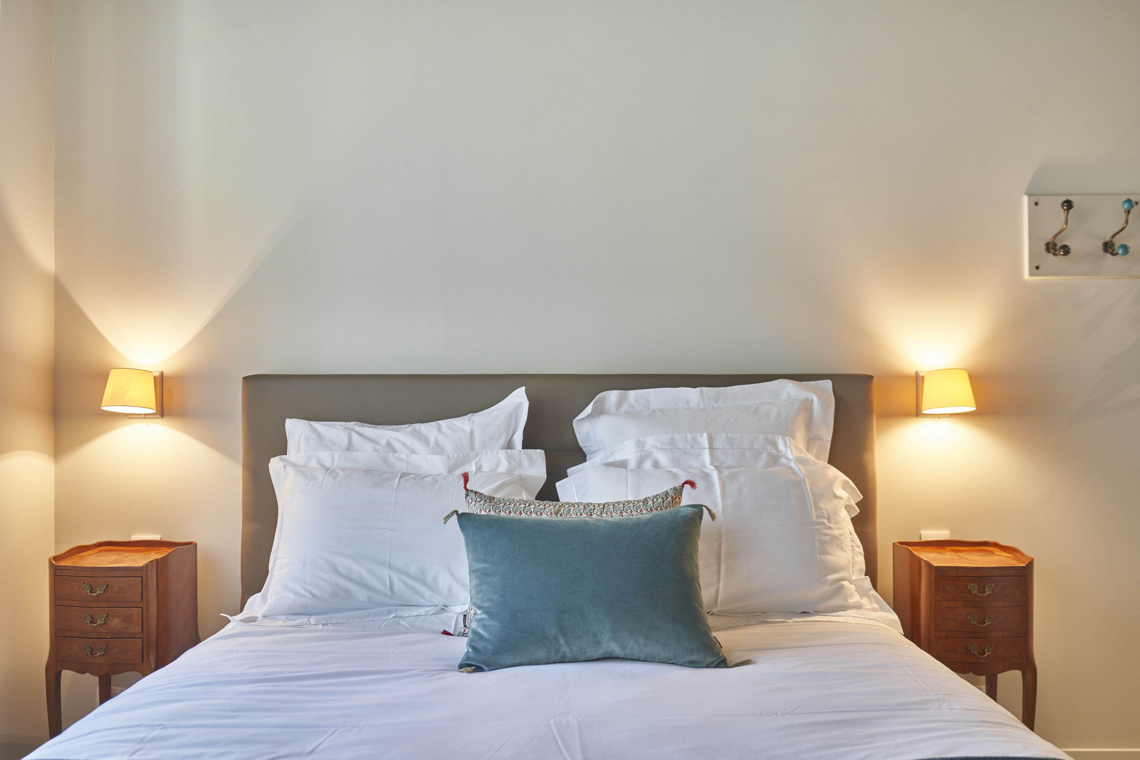 A neatly made bed with white linens and a teal accent pillow, flanked by two bedside tables with lamps, in a simple room with a plain wall and a small picture.