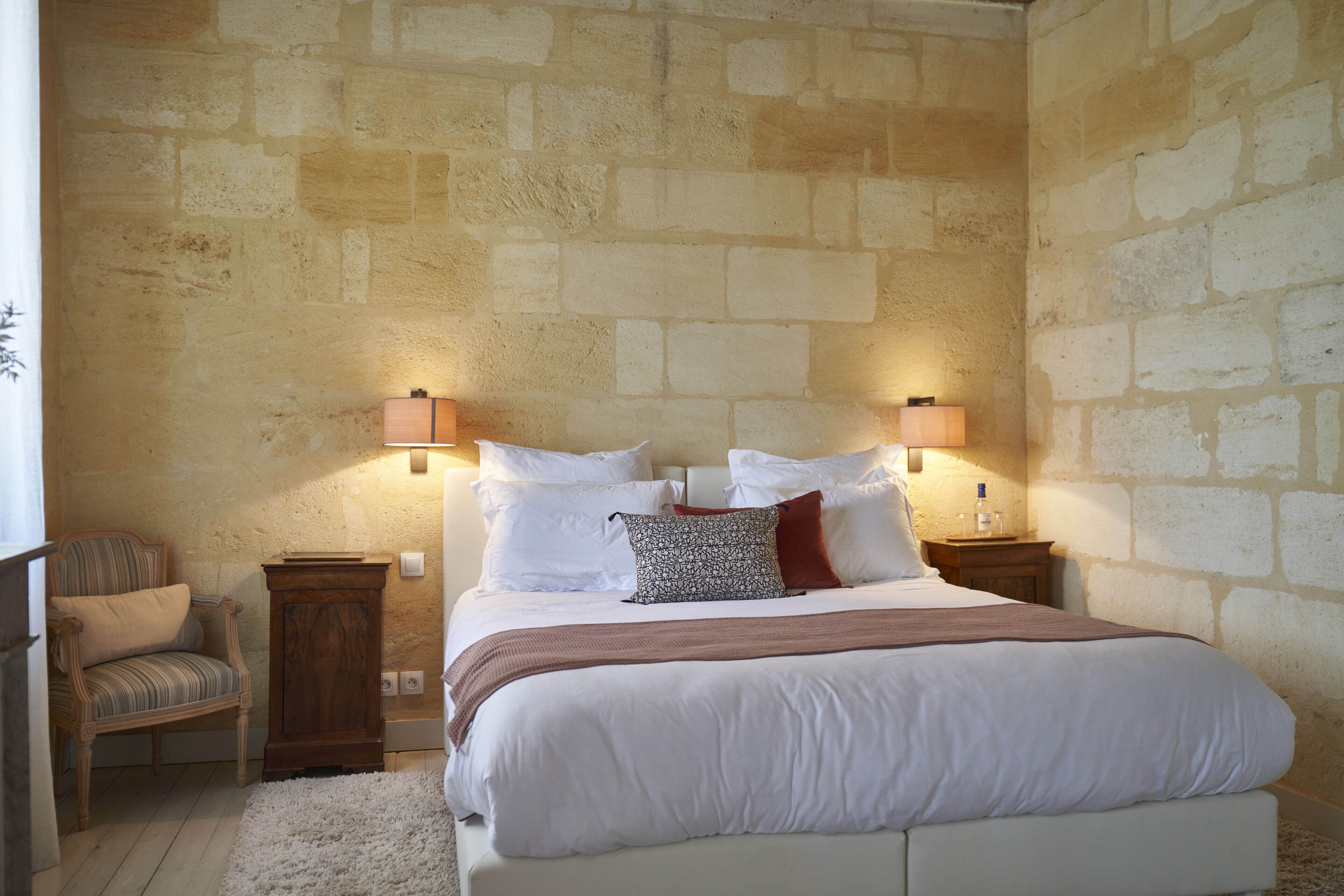 A bedroom with a stone wall, a bed with white bedding, two nightstands, a chair, and a lamp.