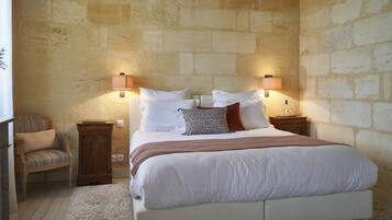 A bedroom with a stone wall, a bed with white bedding, two nightstands, a chair, and a lamp.