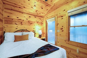 A wooden cabin room with a bed, a nightstand, a lamp, and a window with a view.