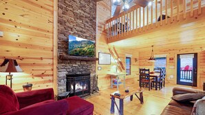 A wooden cabin interior with a stone fireplace, wooden floors, a dining area, and a balcony. - Storybook Hideaway, where memories that fill your heart with comfort, gladness a (Blue Ridge)