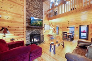 A wooden cabin interior with a stone fireplace, wooden floors, a dining area, and a balcony.