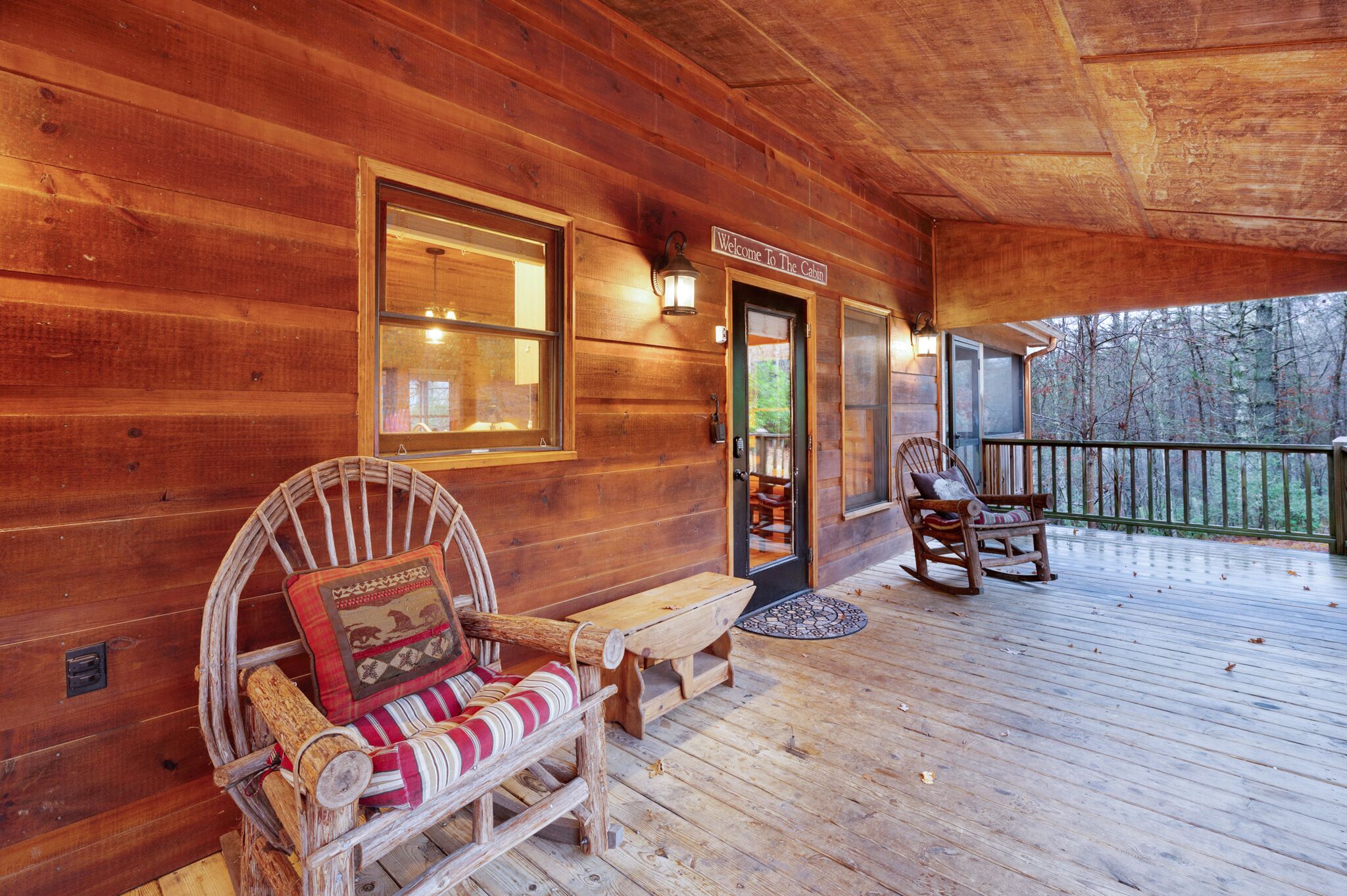 A wooden cabin with a porch, two rocking chairs, a bench, and a door with a 'Welcome To The Cabin' sign.
