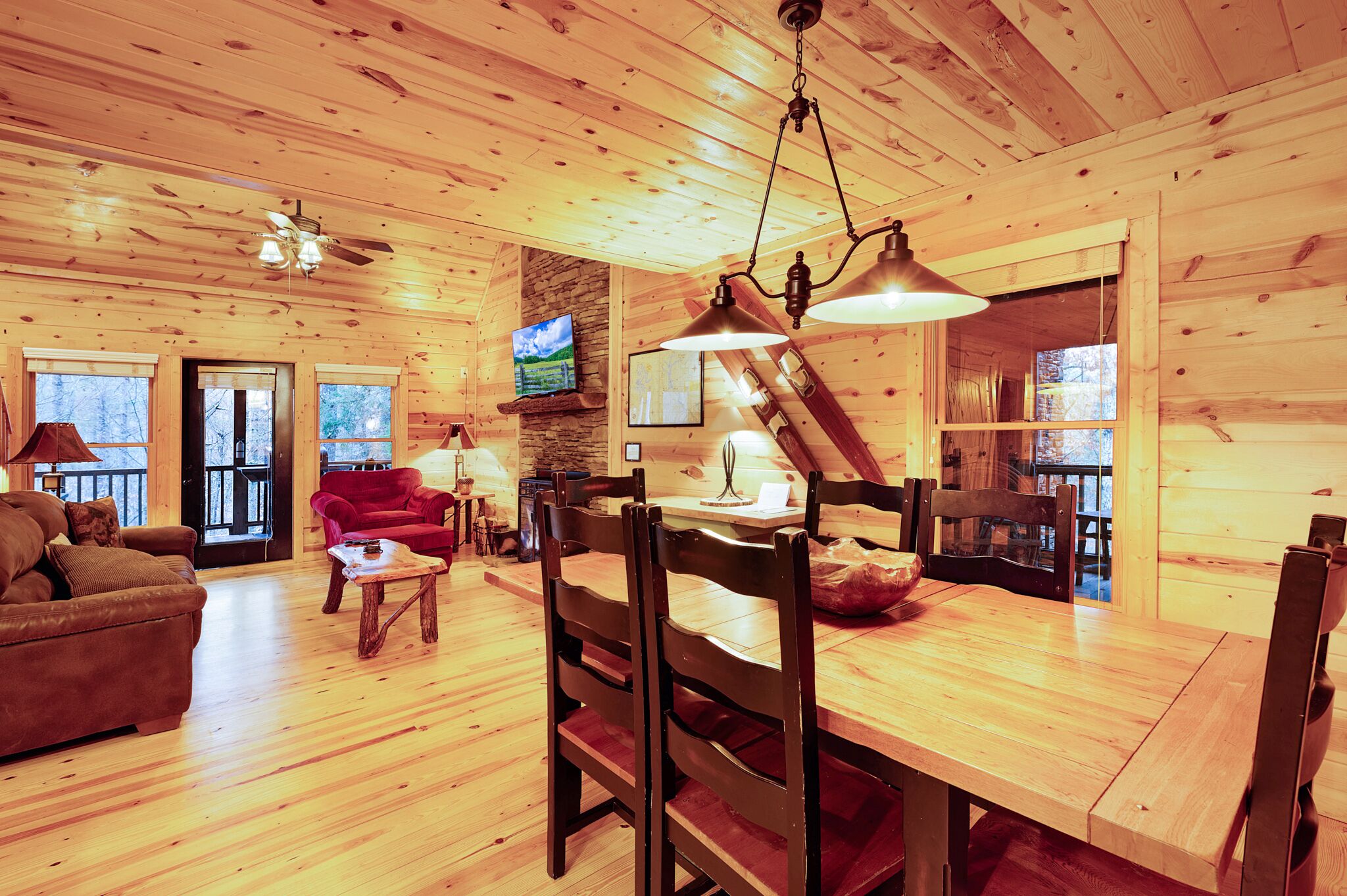 A wooden cabin interior with a dining table, chairs, a red sofa, a television, and a ceiling fan.