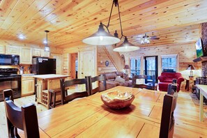 A wooden cabin interior with a dining table, chairs, a kitchen area, and a living room with a couch and fireplace.
