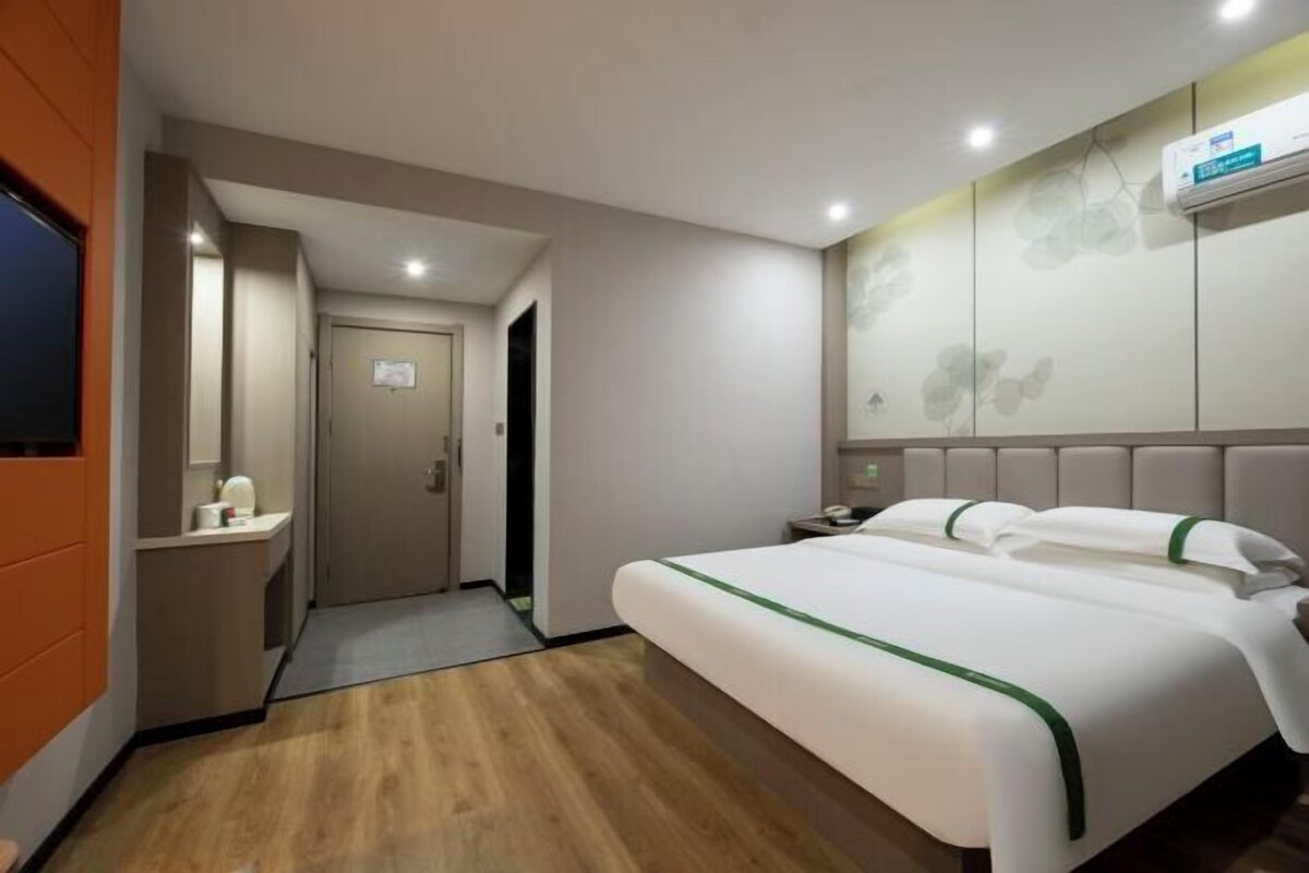A hotel room with a bed, a bedside table, a nightstand, a wall-mounted air conditioner, and a door leading to another room.