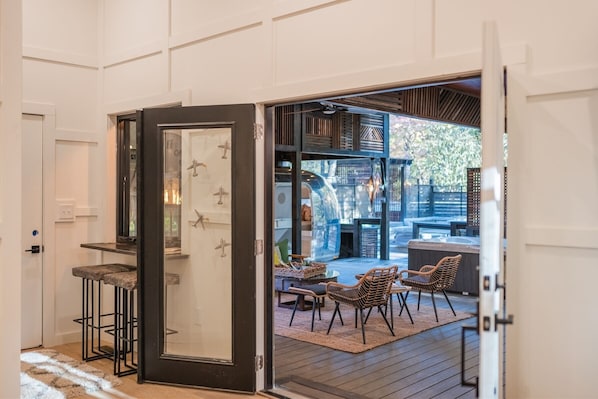 An open door leading to an outdoor patio with wicker furniture, a hot tub, and a view of a backyard with a wooden deck and a hanging light fixture.