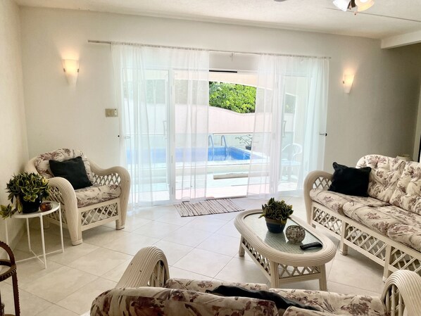 A spacious living room with two wicker sofas, a coffee table, and a potted plant. A sliding glass door leads to a pool area. - Family-Friendly 3BR Villa Apartment w/ Pool – Steps to Surf & Ocean (Green Garden)