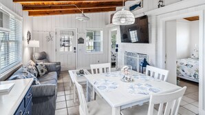 Ein gemütliches Zimmer mit Holzbalkendecke, weißen Wänden, einem Esstisch mit weißem Tischtuch und einer Couch mit gemusterten Kissen.