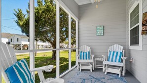 Eine überdachte Veranda mit weißen Adirondack-Stühlen, einer Teppich mit Seestern-Muster und Blick auf einen grünen Garten und Häuser.