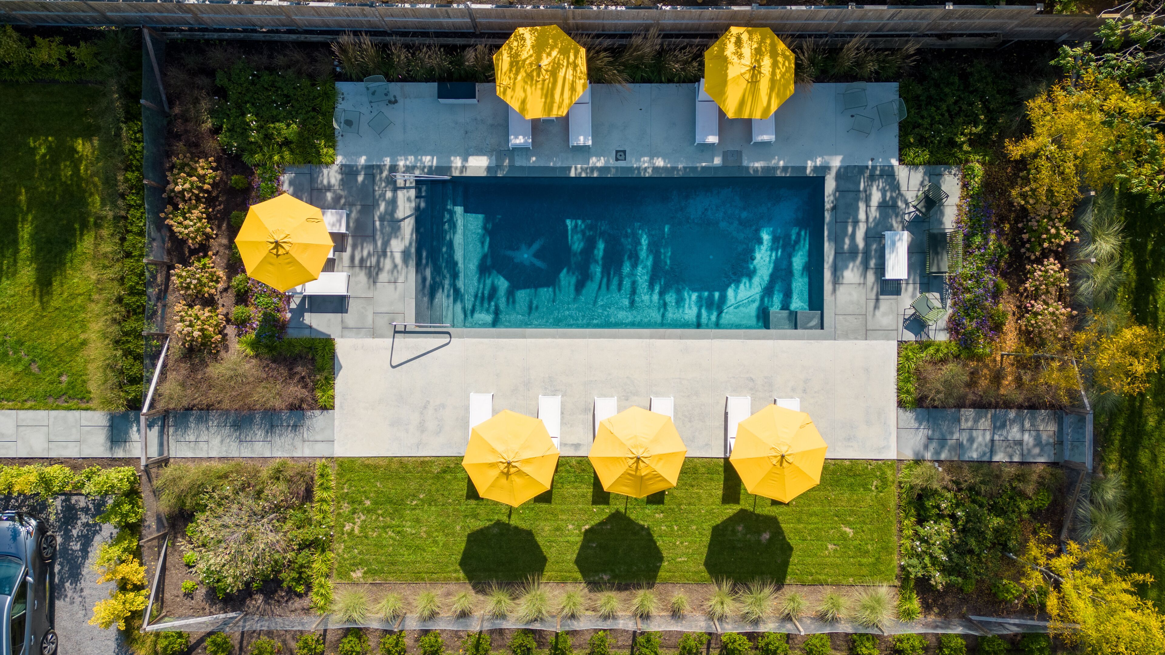 An aerial view of a swimming pool area with multiple yellow umbrellas, a concrete deck, and surrounding greenery.