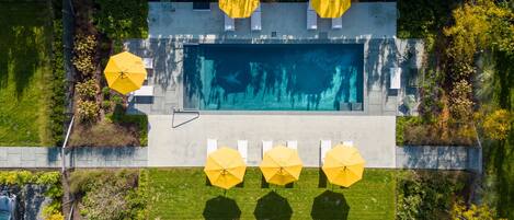 An aerial view of a swimming pool area with multiple yellow umbrellas, a concrete deck, and surrounding greenery.