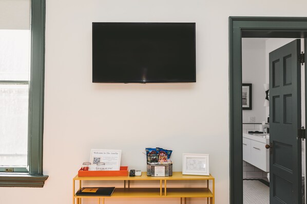 Una habitación con televisor montado, una pequeña mesa con un cartel de bienvenida, aperitivos y un cuadro enmarcado.