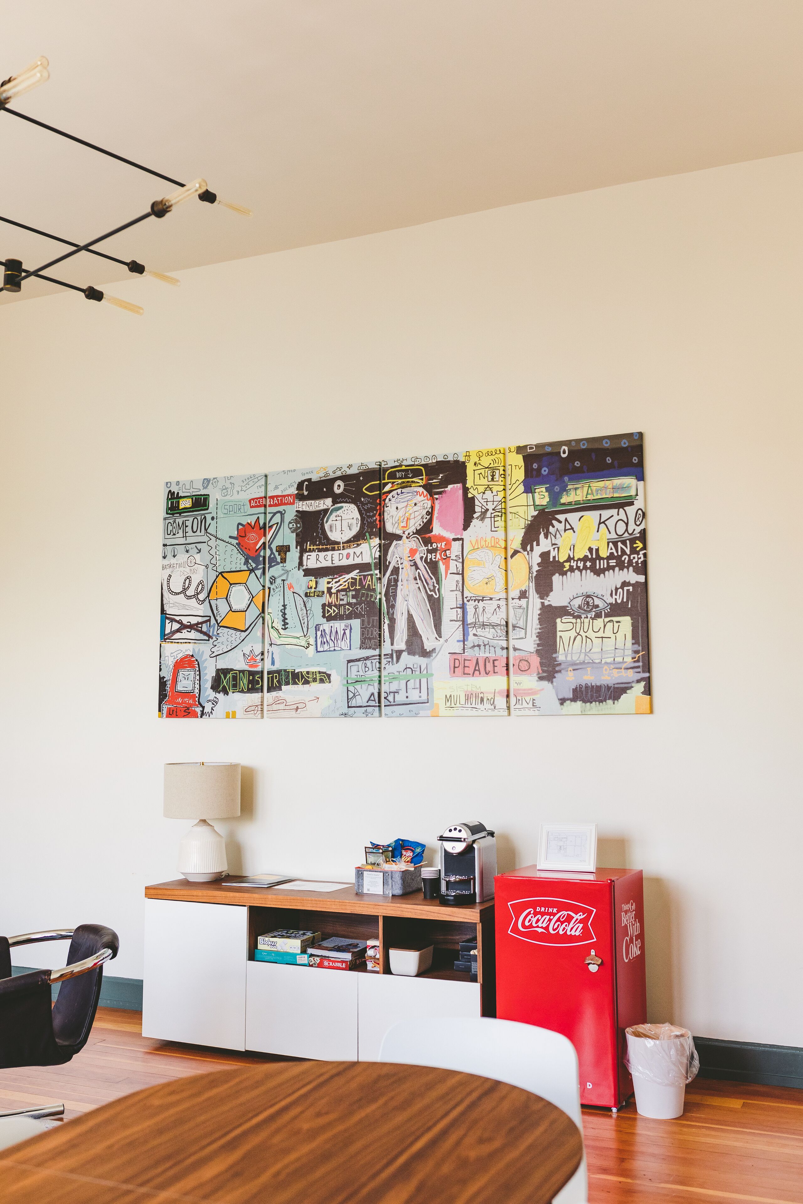 Een kamer met een houten tafel, een witte kast, een rode Coca-Cola koelkast en een muur met een kleurrijke muurschildering.