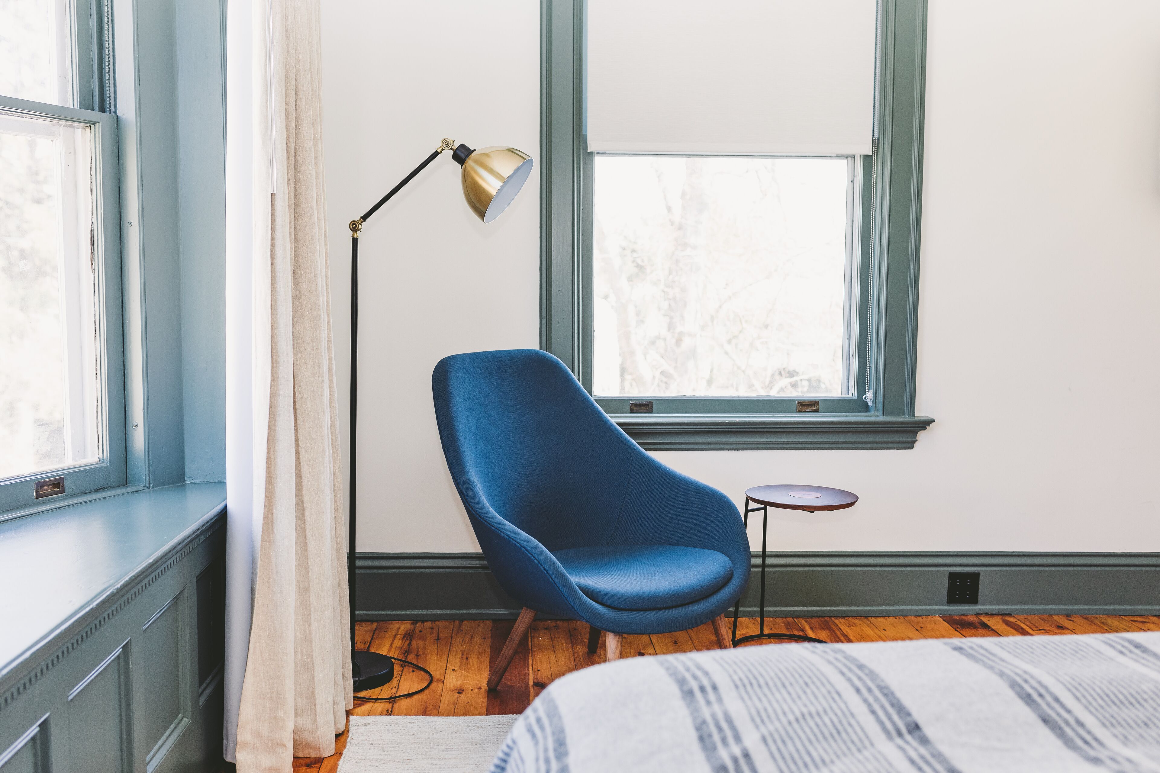 A room with a blue chair, a floor lamp, a small table, and a bed with a striped blanket.