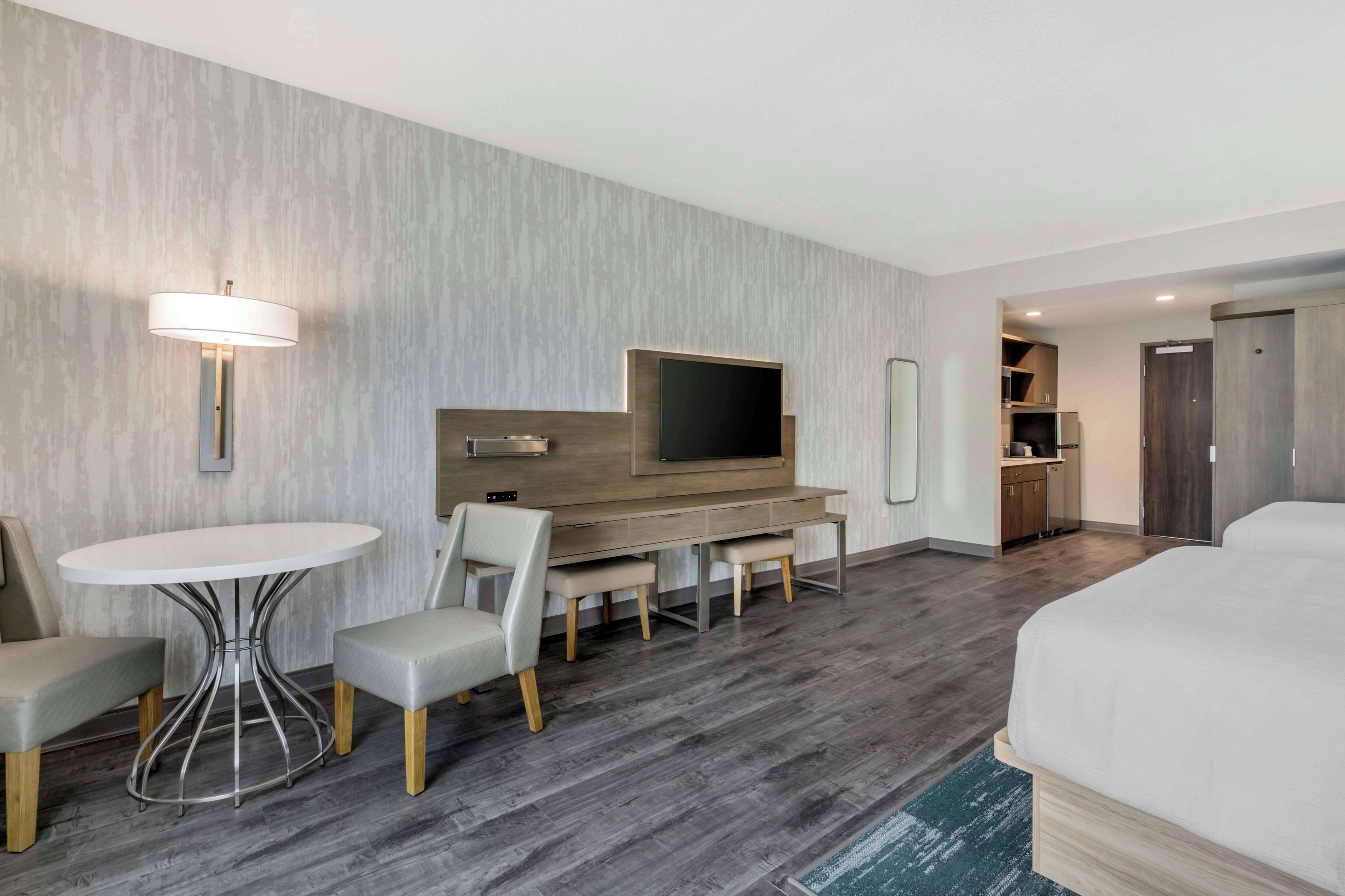 Quarto de hotel com cama, televisão, mesa de jantar com cadeiras, pequena cozinha e banheiro.