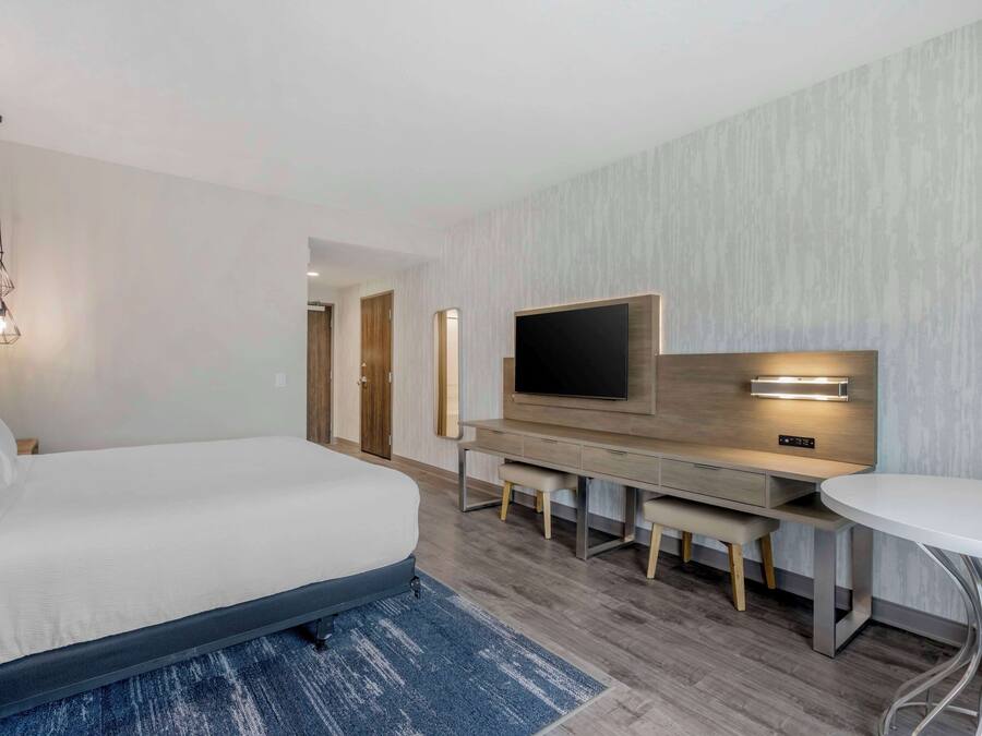 A modern hotel room with a large bed, a desk with a flat-screen TV, and a round dining table.