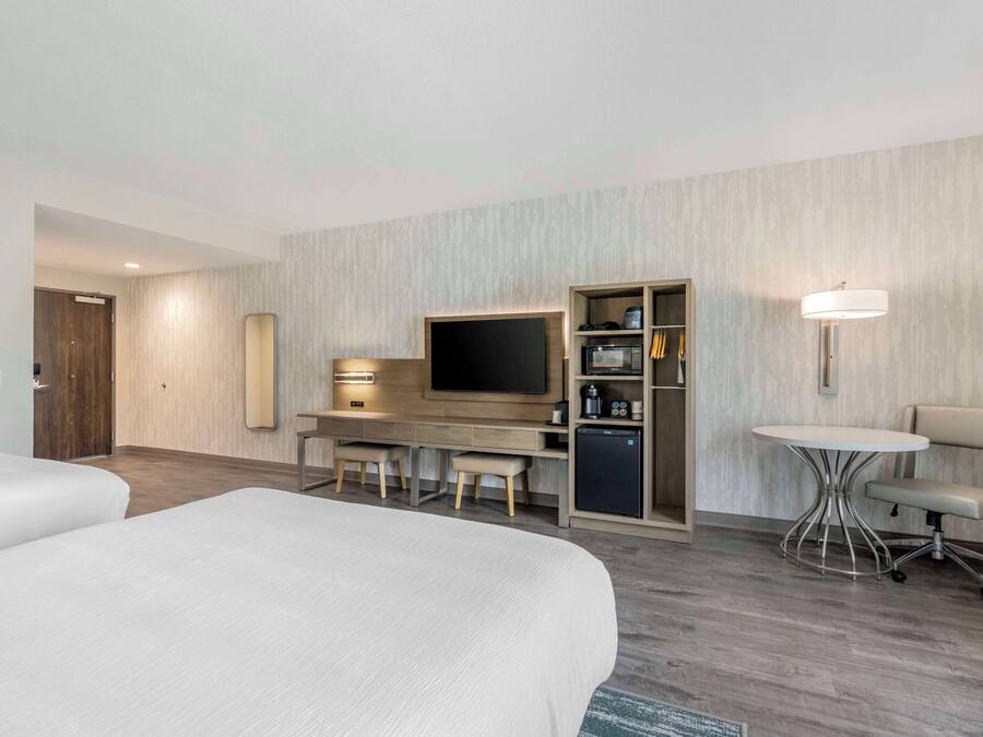 A hotel room with two beds, a desk with a TV, a minibar, and a small table with a chair.
