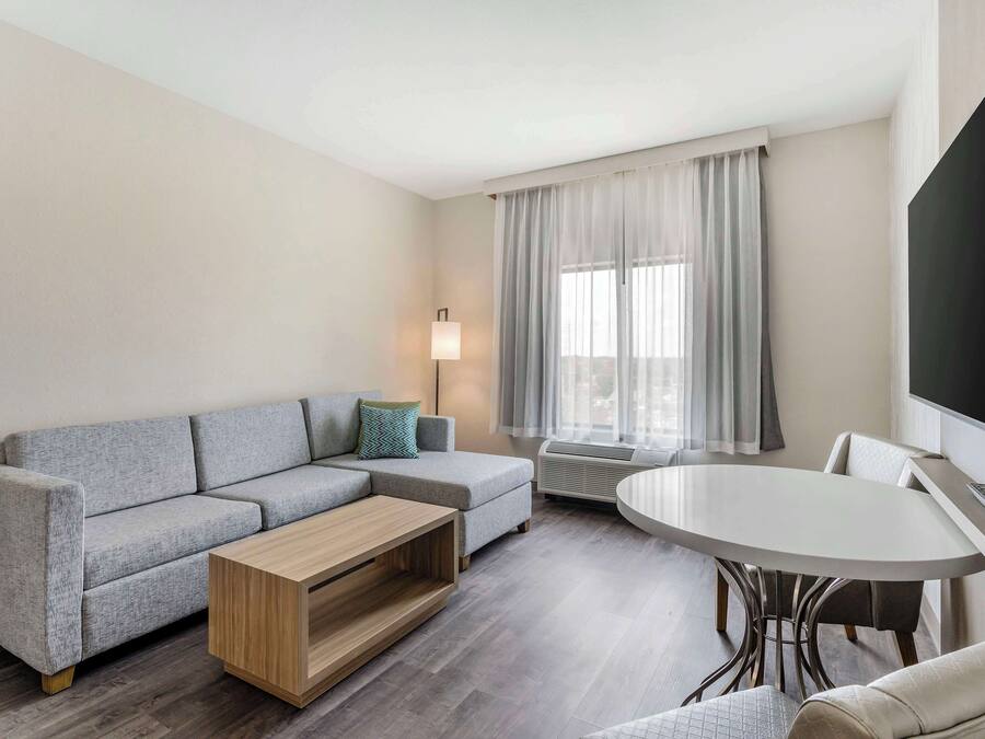 A modern living room with a grey sectional sofa, a wooden coffee table, a round dining table, and a flat-screen TV mounted on the wall.