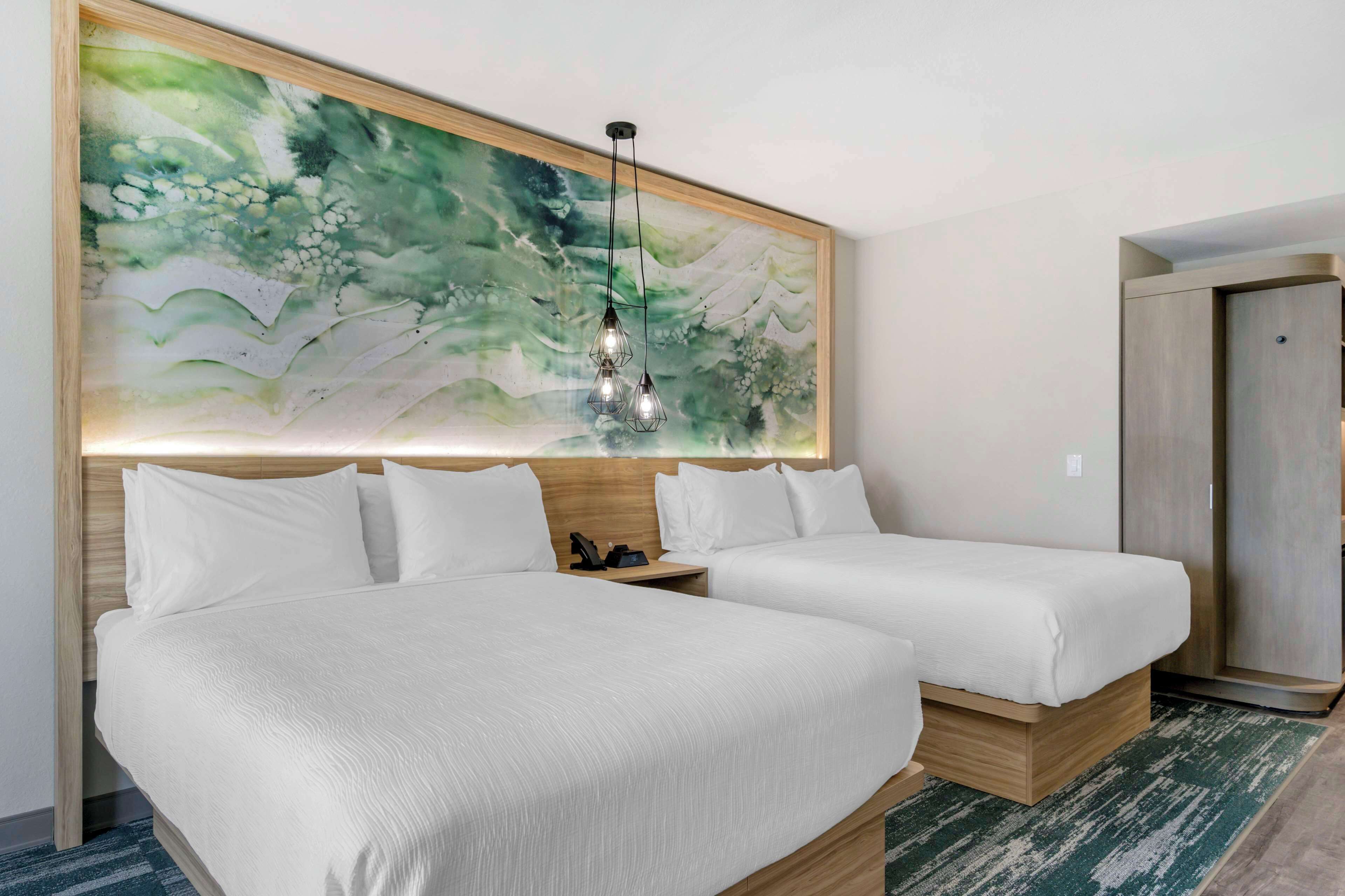Una camera d'hotel con due letti, un quadro astratto di grandi dimensioni appeso al muro, una lampada a sospensione e un armadio in legno.