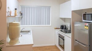 Cocina compacta con armarios blancos, refrigerador de acero inoxidable, microondas, pileta y ventana con persianas.