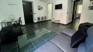 A modern living room with a glass coffee table, a grey sofa, and a TV mounted on the wall. There are three stools and a small table with a candle on the floor. - Bonaventura s House by Wonderful Italy (Olbia)
