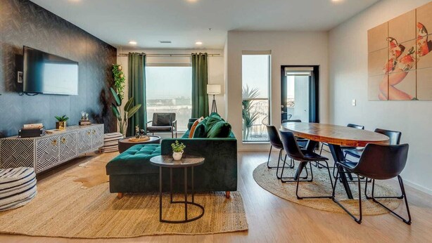 A modern living room with a sofa, coffee table, and TV. There is a dining area with a table and chairs. The room has large windows and a view of the city.